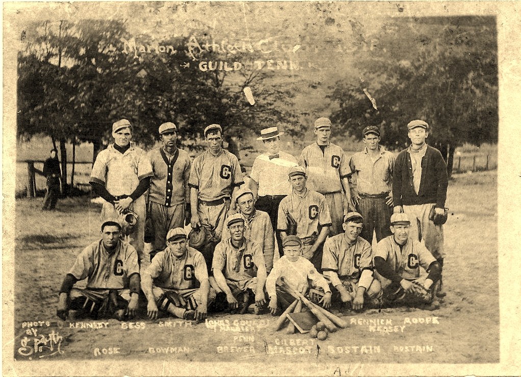Team photograph of fourteen men and a boy in baseball uniforms posed outdoors, with players' surnames lettered across the bottom of the print